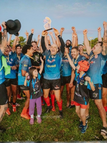Fotografia colorida em plano médio de uma equipe mista de rúgbi, com cerca de 12 atletas em uniformes azuis e pretos, comemorando em um gramado. Três crianças também vestindo as cores do time estão na frente, misturadas com os adultos. O grupo está aglomerado e sorrindo, e ao centro, uma jogadora segura um troféu prateado no ar com as duas mãos, celebrando a conquista.