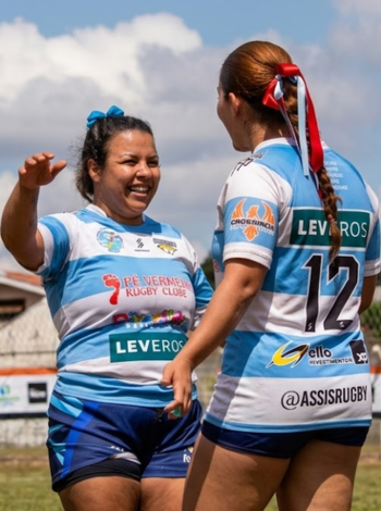 Fotografia de duas jogadoras de rúgbi sorrindo e interagindo em um campo gramado durante o dia. Elas vestem o uniforme oficial da união Pé Vermelho, com listras horizontais em azul claro e branco e detalhes em azul marinho. No uniforme, é possível ver o logotipo da patrocinadora Leveros em destaque. Uma das atletas está de costas, exibindo o número 12, enquanto a outra está de frente, sorrindo e com a mão estendida em um gesto de celebração ou conversa.