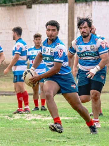 Jogador de rugby do time Assis Rugby correndo com a bola durante treinamento em campo de grama. Ele veste uniforme listrado em azul e branco. Outros jogadores estão ao fundo.
